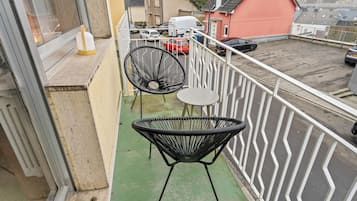 Ein Balkon mit zwei schwarzen Korbstühlen und einem kleinen Tisch, der auf einen Parkplatz mit mehreren Autos und einem Gebäude mit roter Fassade blickt.