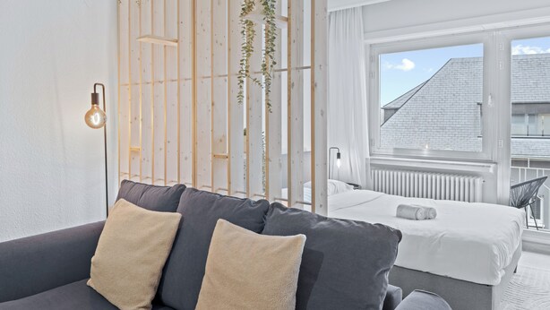A modern hotel room with a grey sofa, a white bed, and a view of a building through large windows.
