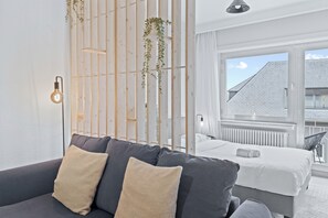 A modern hotel room with a grey sofa, a white bed, and a view of a building through large windows.
