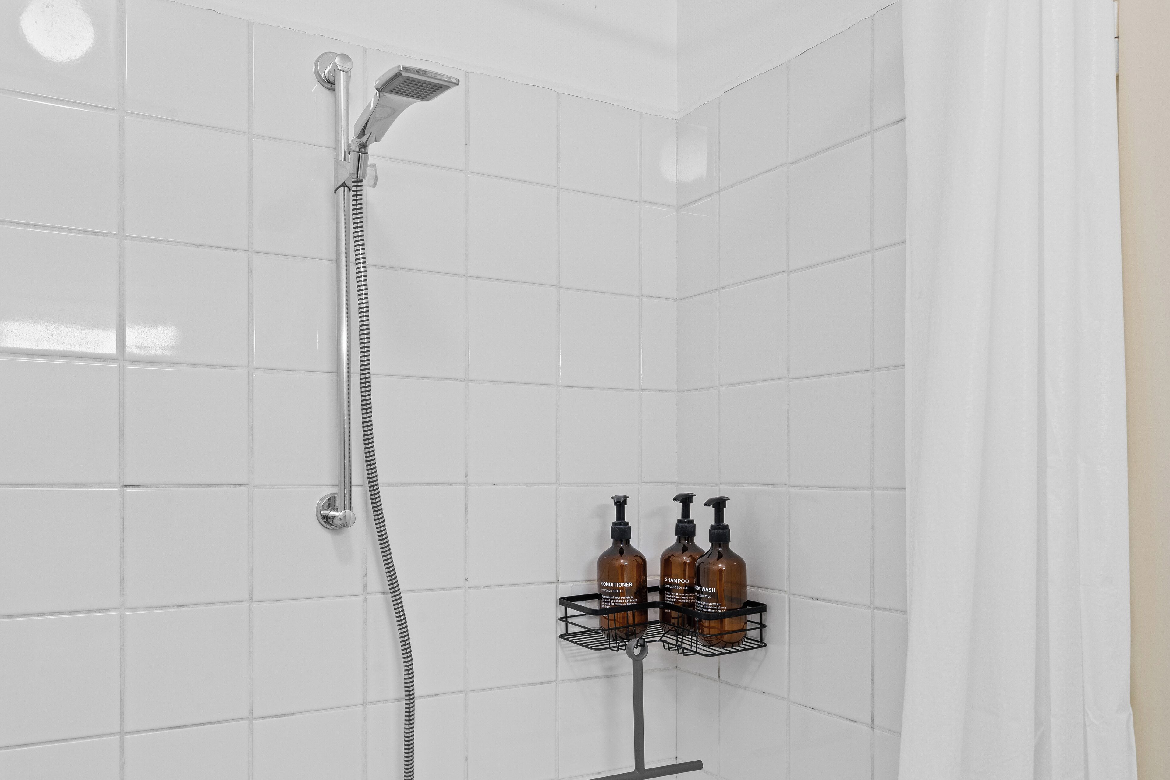 Ein Duschbereich mit weißen Fliesen, einem Handbrausekopf und einem Metallregal mit drei Shampoo- und Conditionerflaschen.