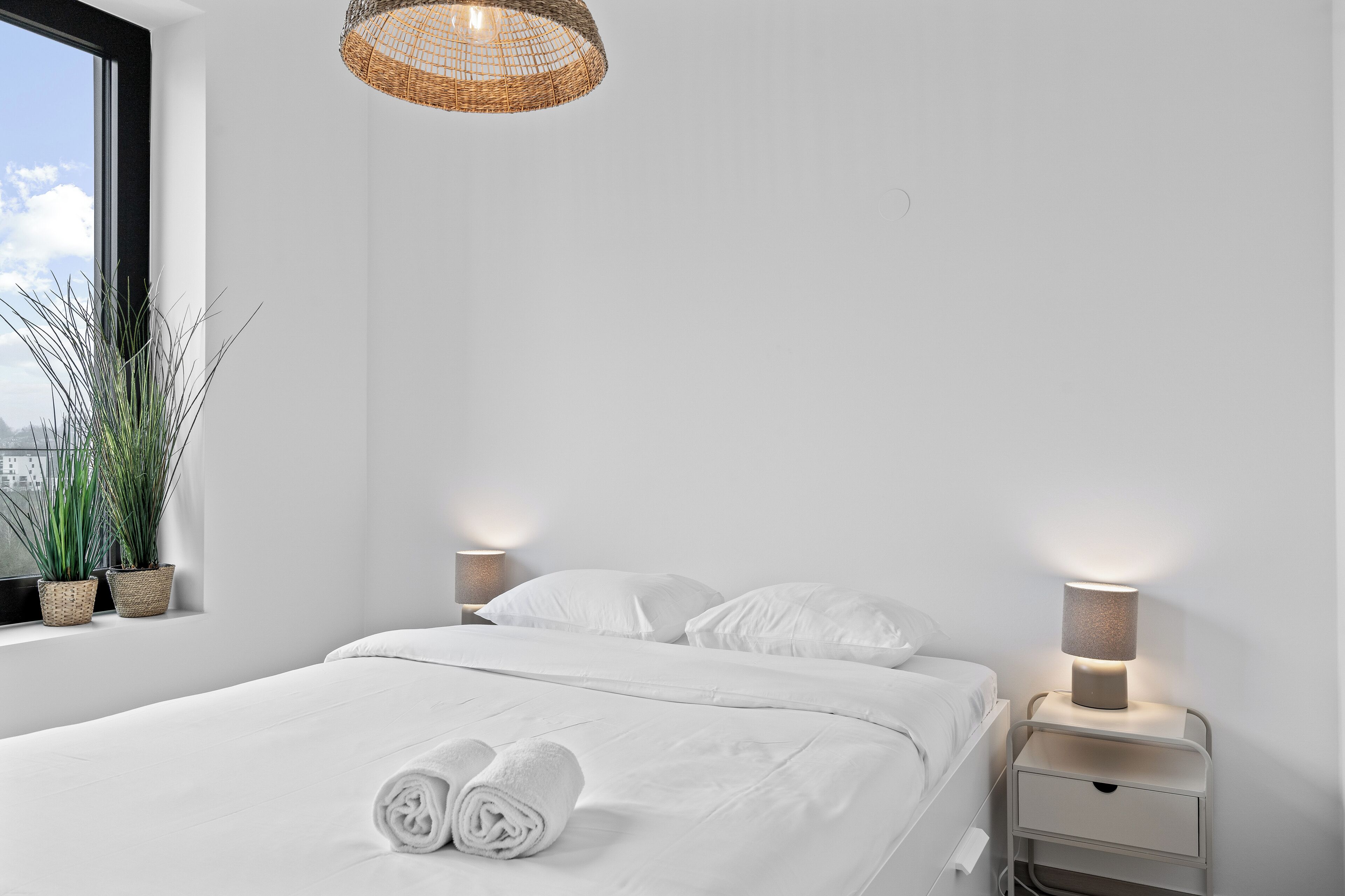 A neatly made bed with white linens and two rolled towels, a bedside lamp, and a wicker ceiling light fixture in a modern, minimalistic room with a large window.