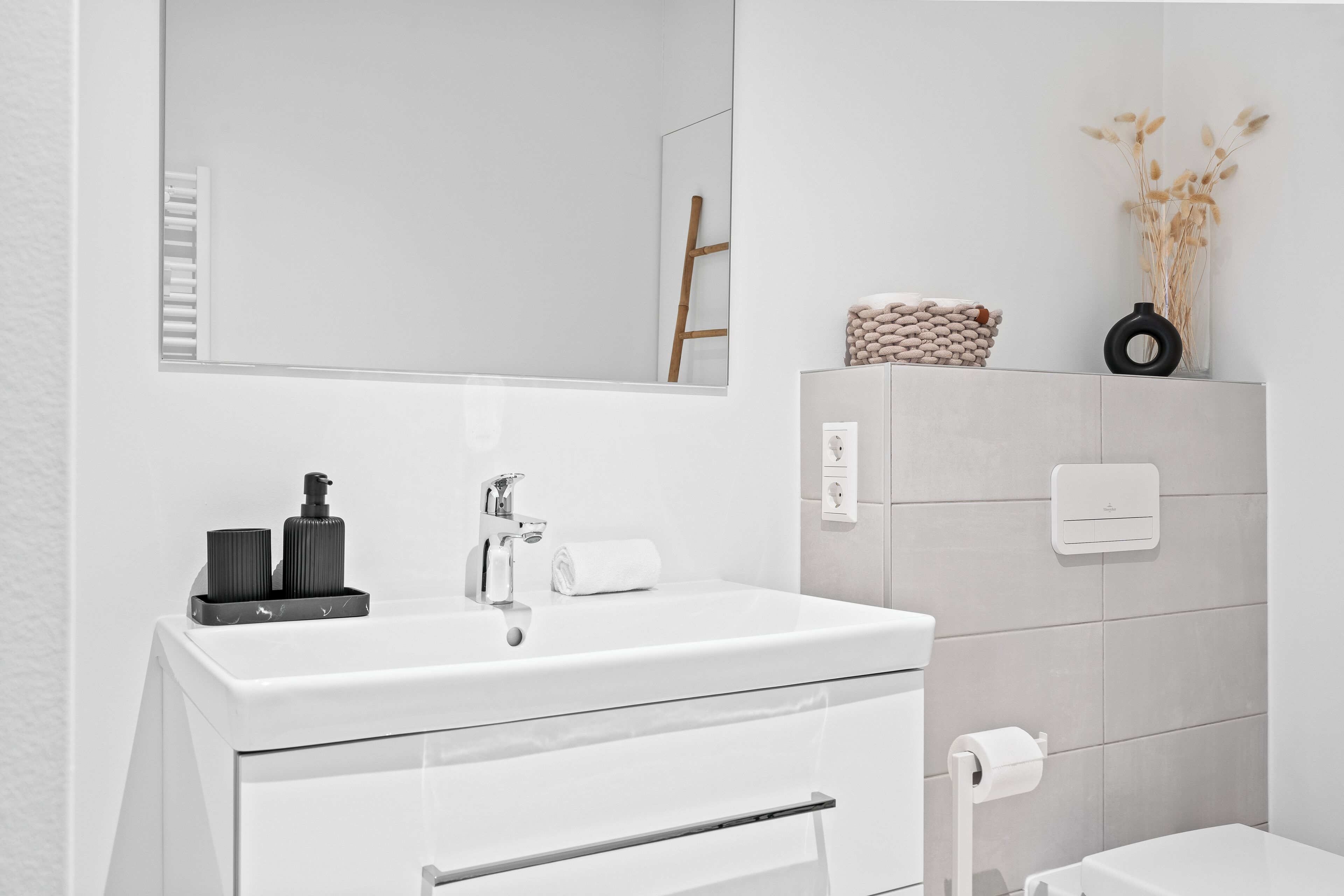 Ein modernes Badezimmer mit einem weißen Waschbecken, Spiegel und Toilette. Es gibt einen Handtuchhalter, eine Toilettenpapierhalterung und ein kleines Regal mit Dekorationen.