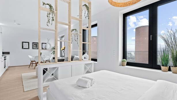 A modern hotel room with a large bed, a wooden desk, a white chair, and a view of the city through large windows.