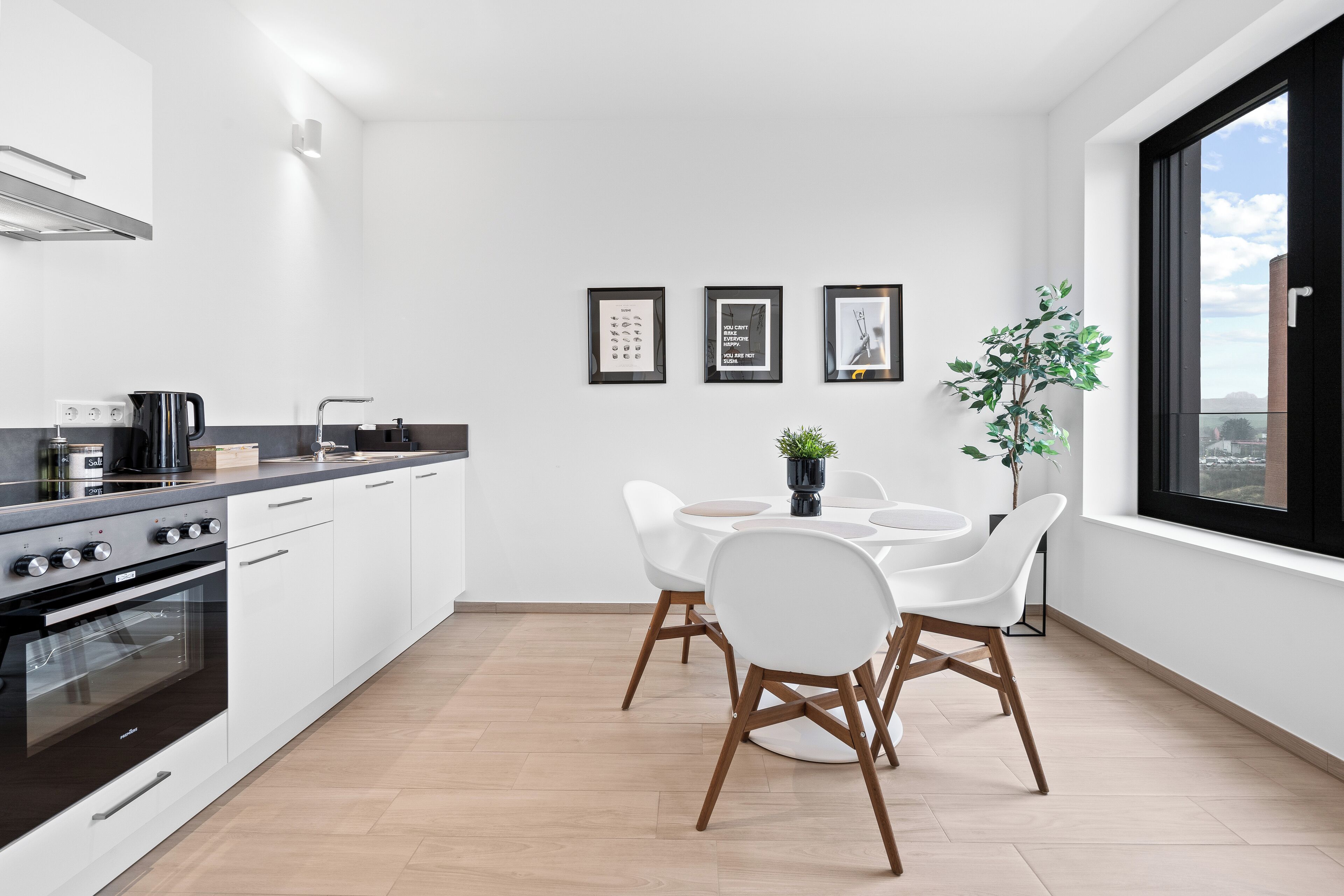 A modern kitchen with white cabinets, a black stove, and a dining area with white chairs and a round table. There are three framed pictures on the wall and a potted plant near the window.