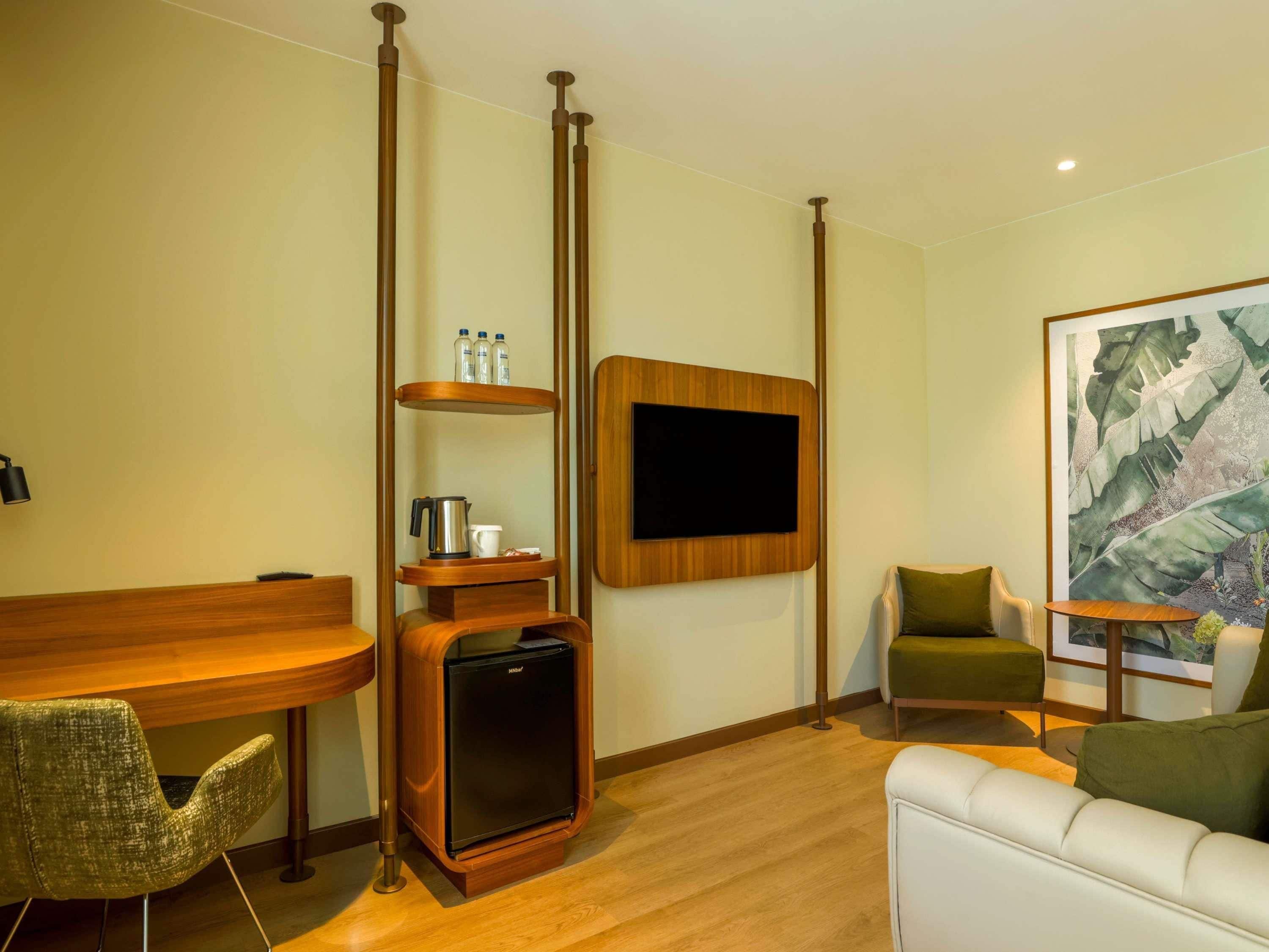 A modern hotel room with a wooden desk, a small refrigerator, a flat-screen TV mounted on the wall, a green armchair, a white sofa, and a large abstract painting.