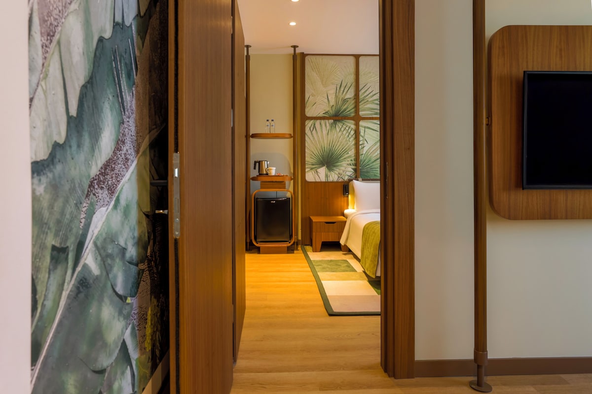 A hotel room with a wooden floor, a bed with a green blanket, a bedside table with a lamp, a TV mounted on the wall, and a large window with a plant.