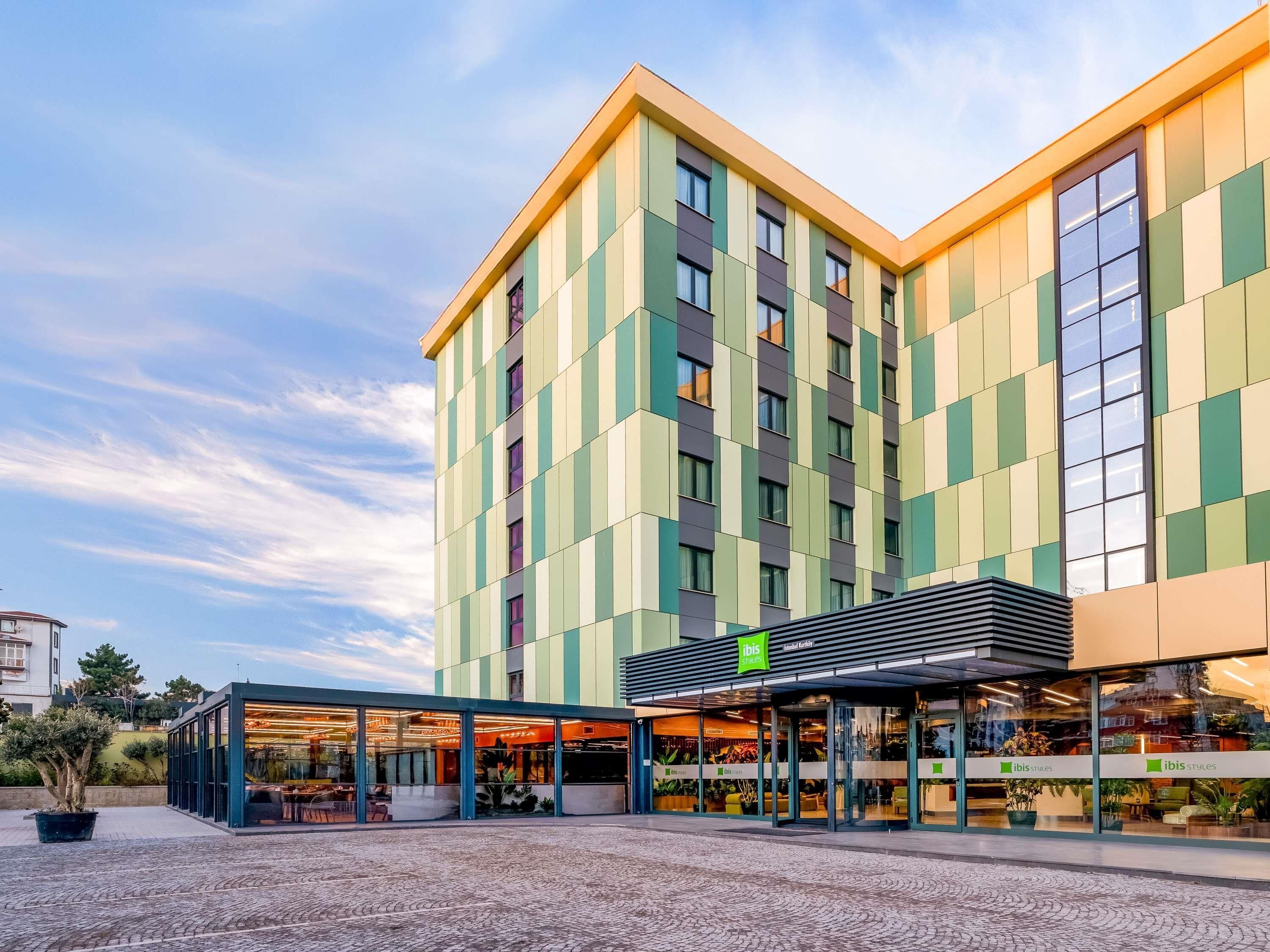 A modern hotel with a green and beige facade, large windows, and a covered outdoor seating area.