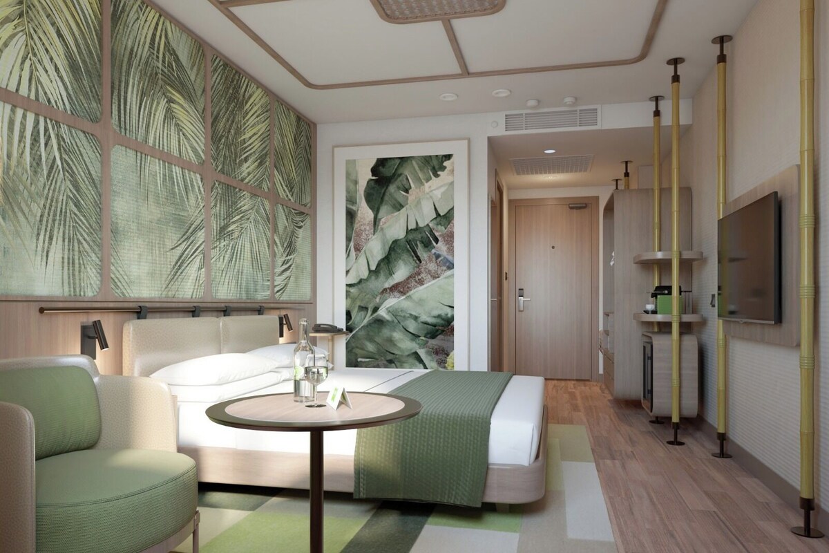 A modern hotel room with a bed, a round table with a bottle and glass, a green armchair, a large wall mural, and a TV mounted on the wall.