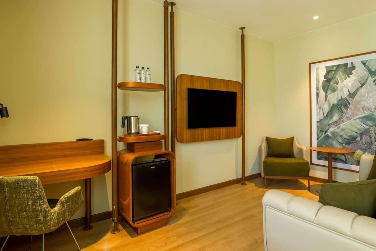 Habitación de hotel moderna con un escritorio de madera, un refrigerador pequeño, un televisor de pantalla plana montado en la pared, un sillón verde, un sofá blanco y un cuadro abstracto de gran tamaño.