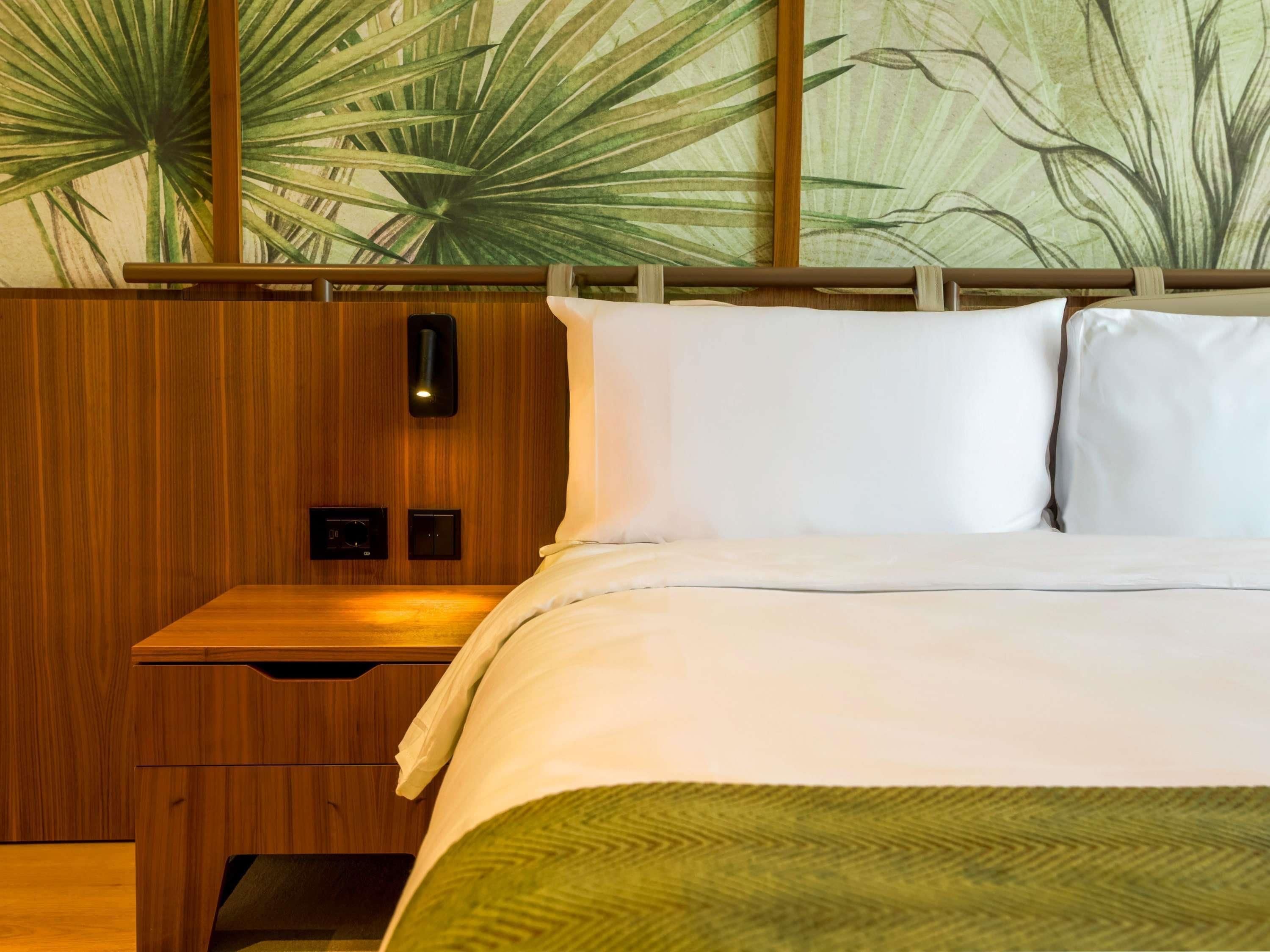A hotel room with a wooden headboard, a bed with white linens, a nightstand with a drawer, and a wall with a large plant mural.