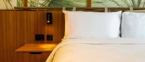 A hotel room with a wooden headboard, a bed with white linens, a nightstand with a drawer, and a wall with a large plant mural.