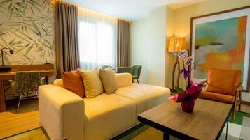 Una habitación de hotel con sofá, una mesita de madera, escritorio con lámpara, una silla y un cuadro en la pared.