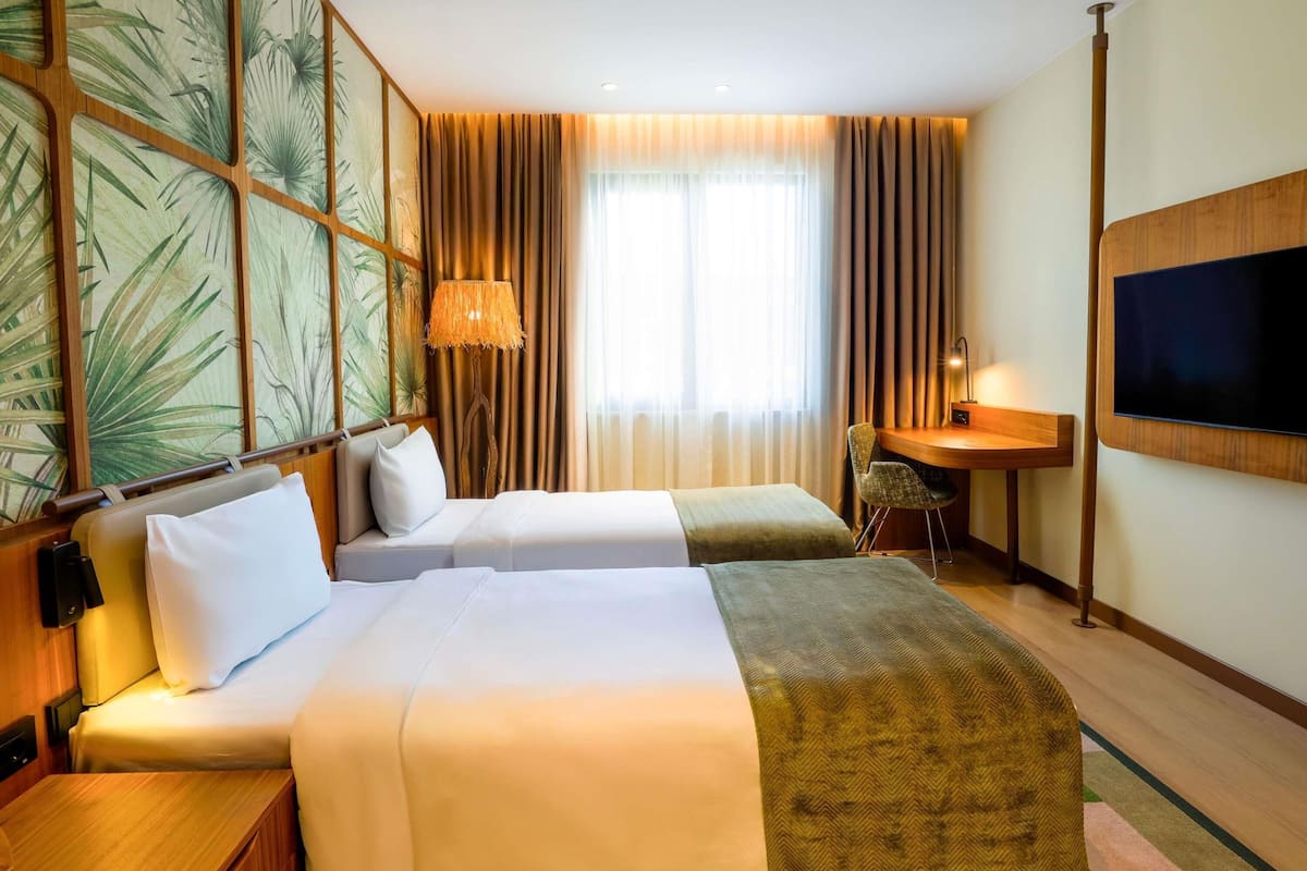 A hotel room with two beds, a desk, a TV, and a large window with curtains.