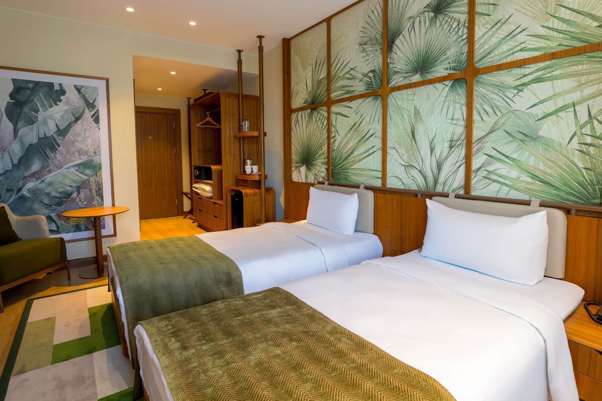 A hotel room with two beds, a wooden wardrobe, a small table, and a large botanical wall mural.