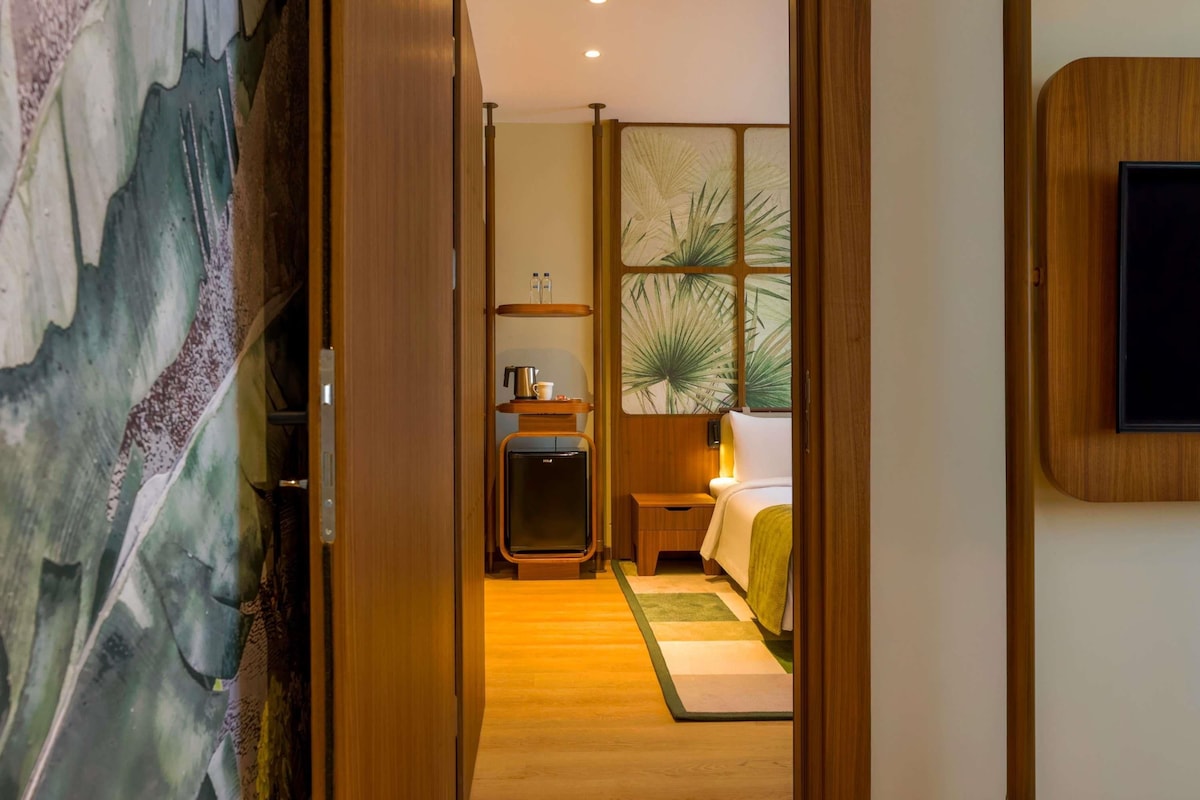 A hotel room with a wooden floor, a bed with a green blanket, a bedside table with a lamp, a small wooden cabinet, and a large window with a plant view.