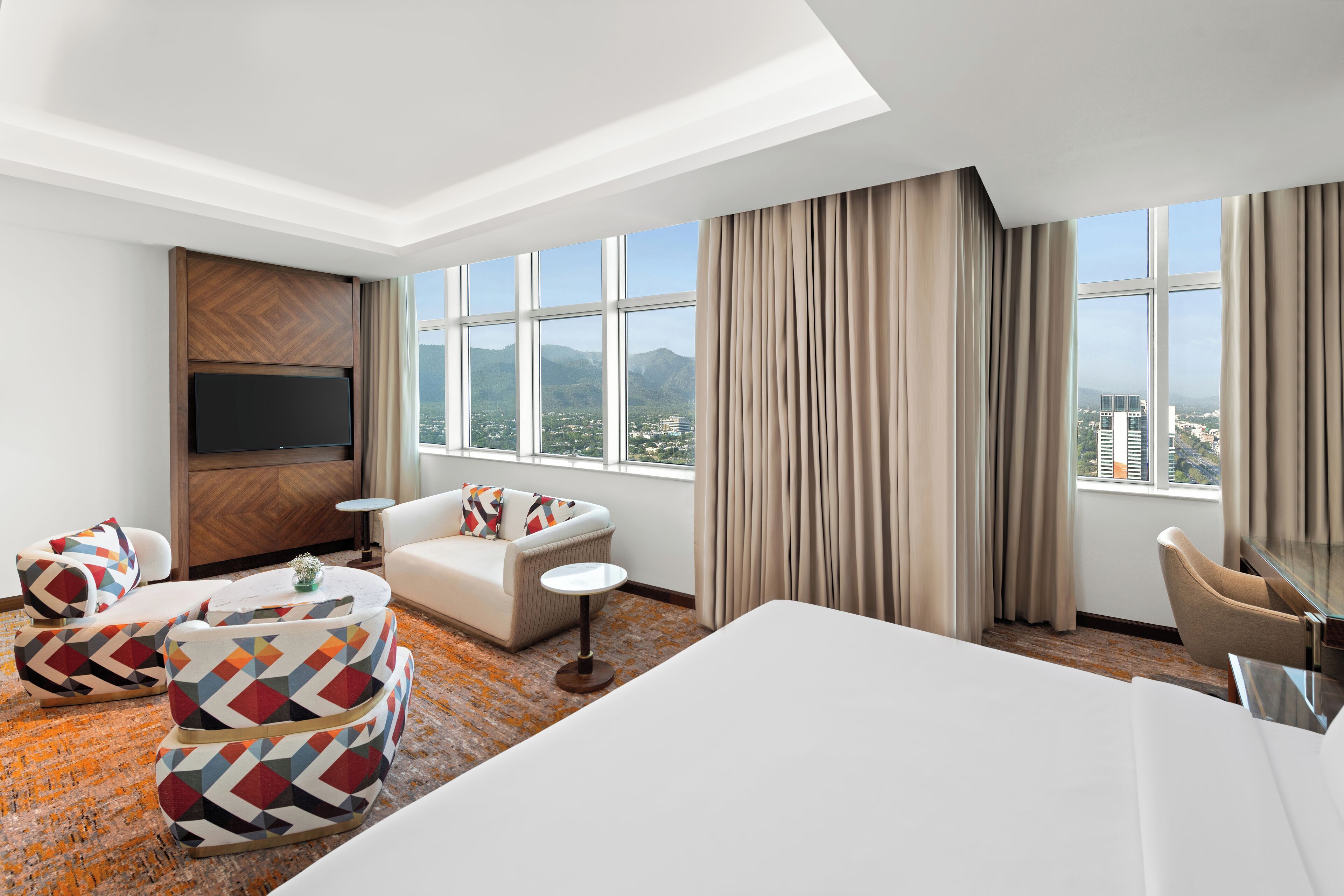 Ein Hotelzimmer mit einem großen Bett, zwei Sesseln, einer Couch, einem kleinen runden Tisch und einem Fernseher. Das Zimmer verfügt über große Fenster mit Blick auf Berge und Gebäude.