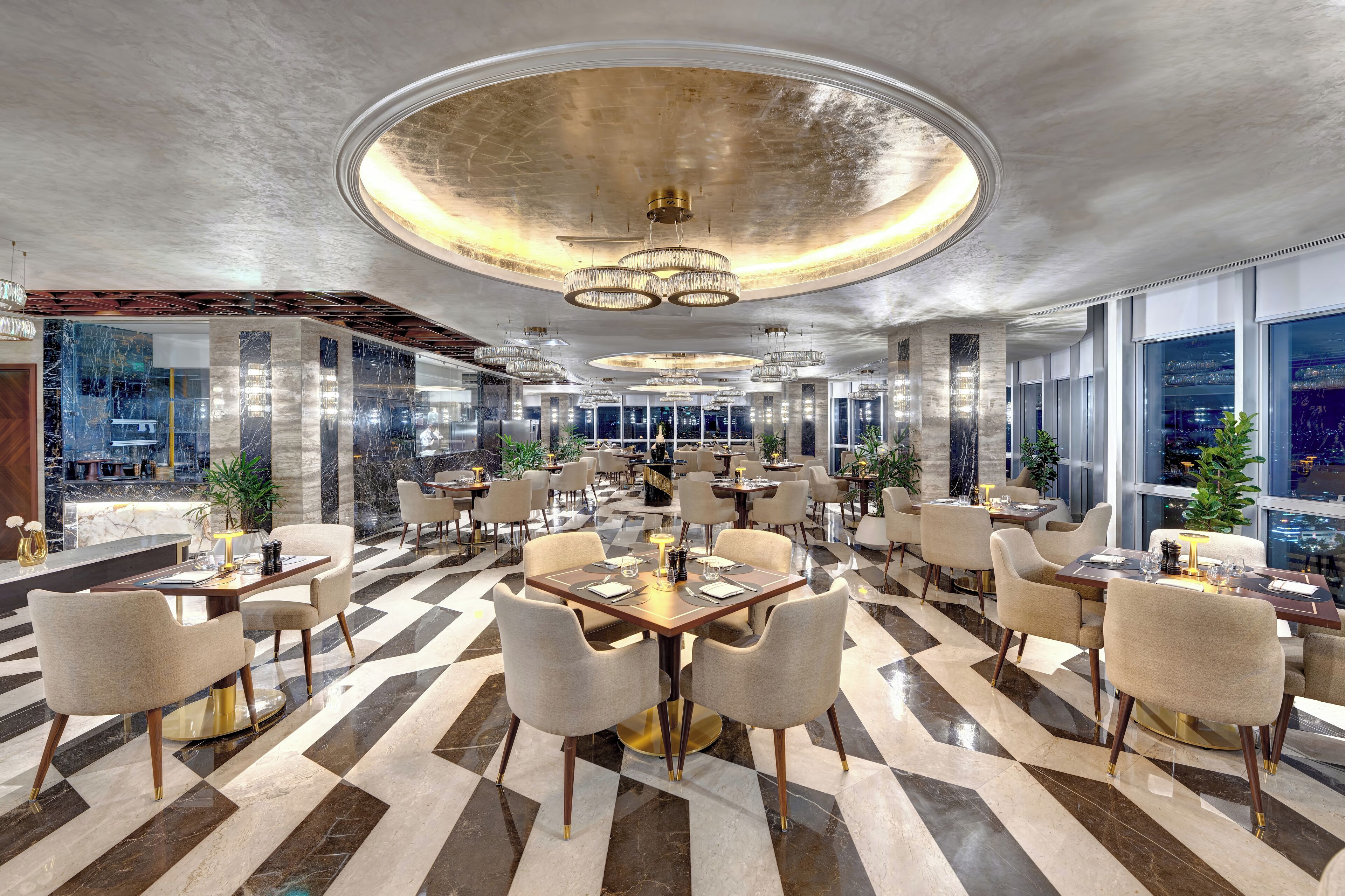 A spacious hotel lobby with a checkered floor pattern, multiple dining tables, and modern chandeliers.