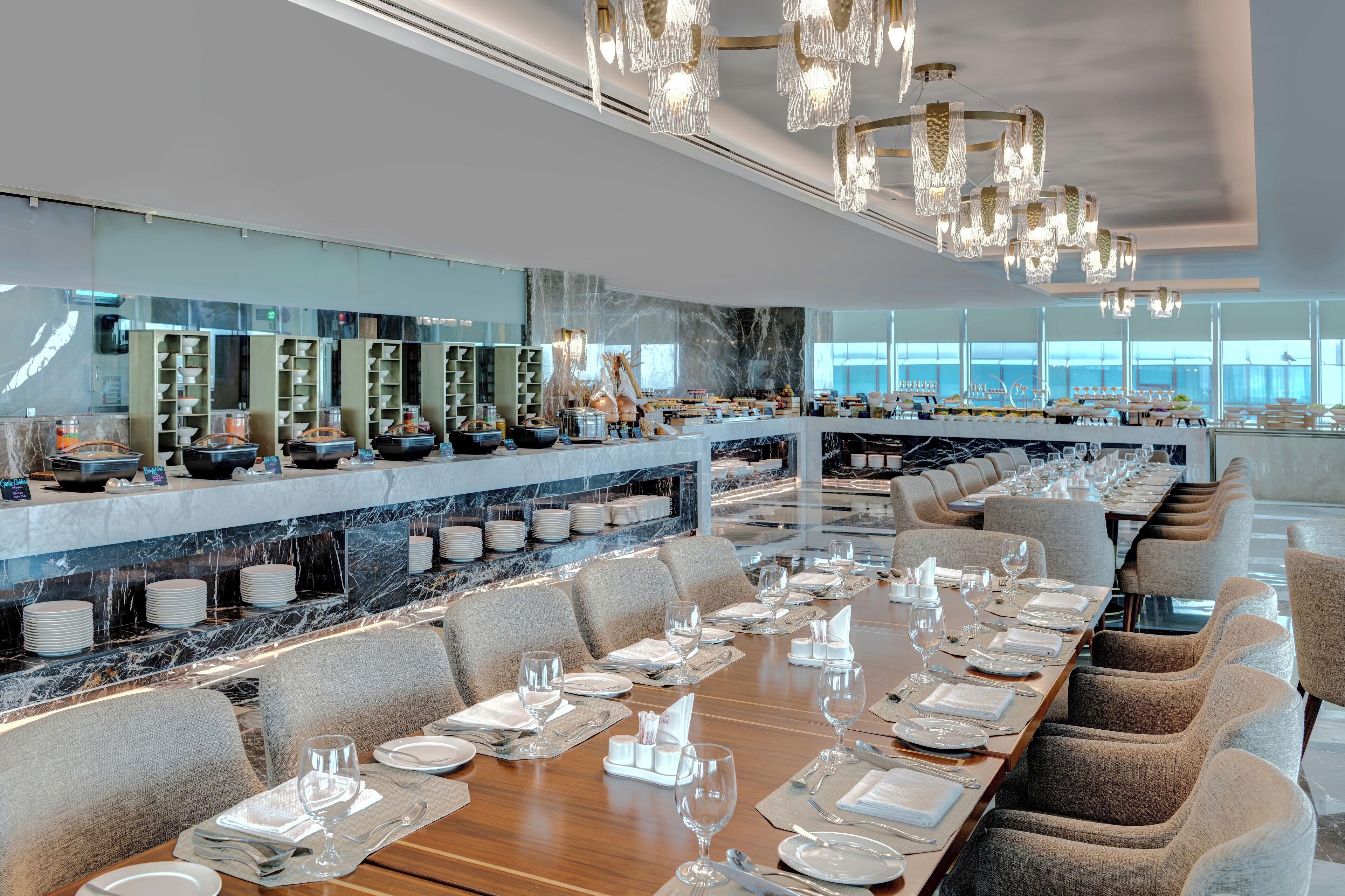 A modern restaurant with a long dining table set for a meal, a marble bar, and a view of the city.
