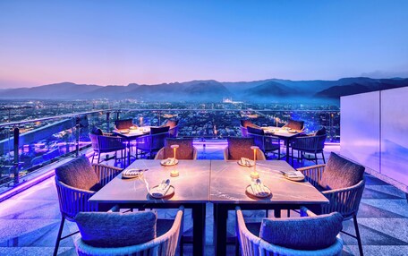 A rooftop dining area with tables and chairs, overlooking a cityscape and mountains.