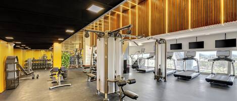 Un gimnasio con diverso equipo de ejercicio, incluyendo cintas de correr y máquinas de pesas, y amplias ventanas que ofrecen vistas de la ciudad.