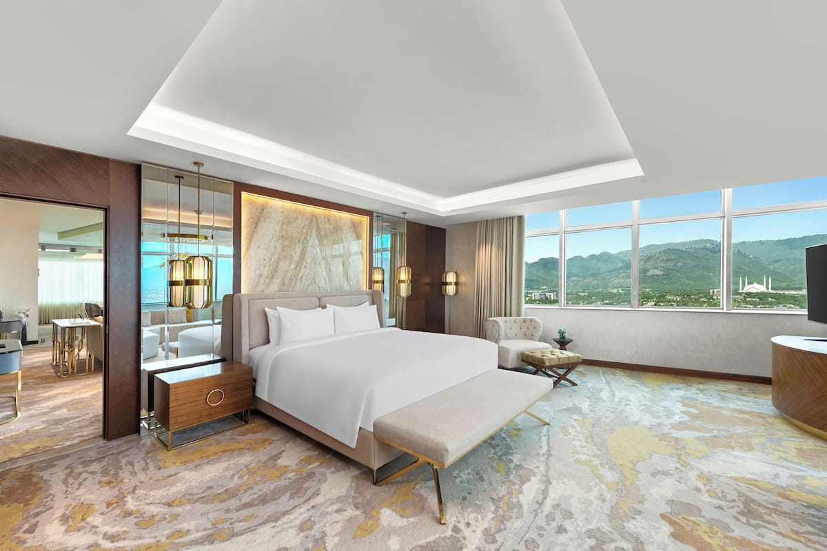 A modern hotel room with a large bed, a bench, a desk, and a view of the mountains.