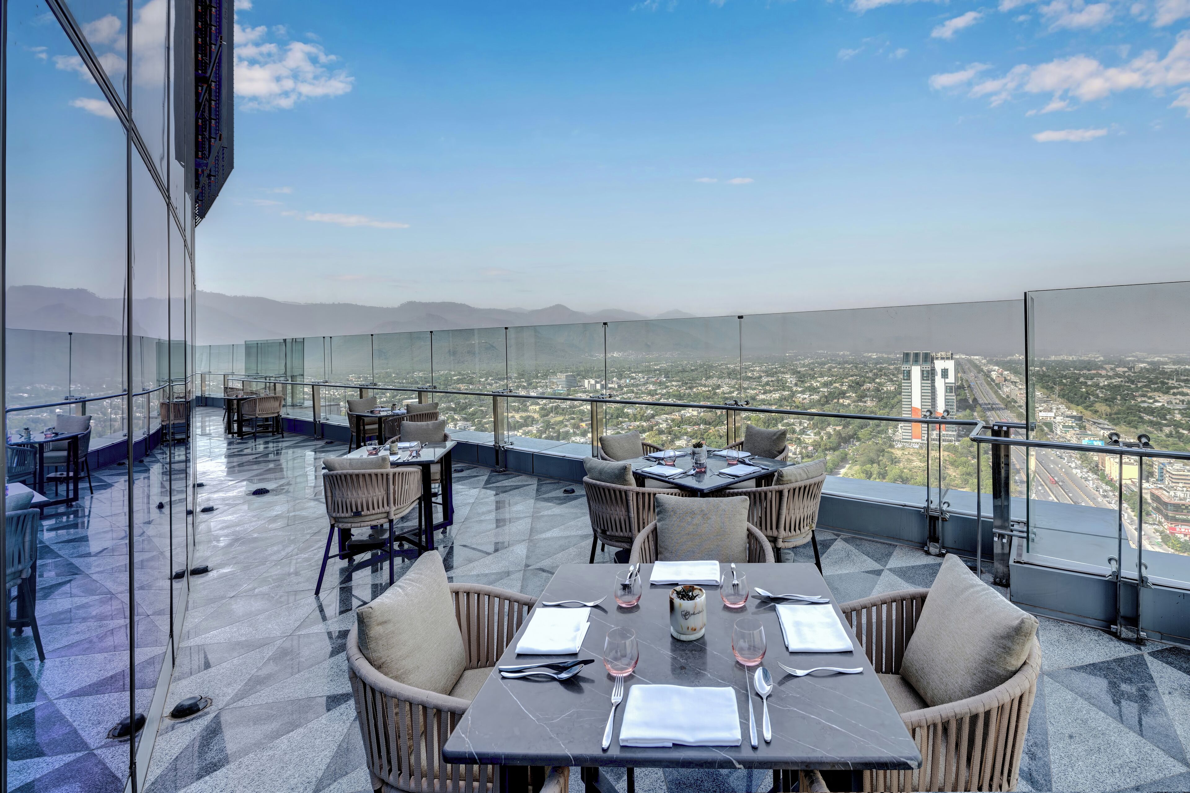 A rooftop dining area with tables and chairs, a glass railing, and a city view.