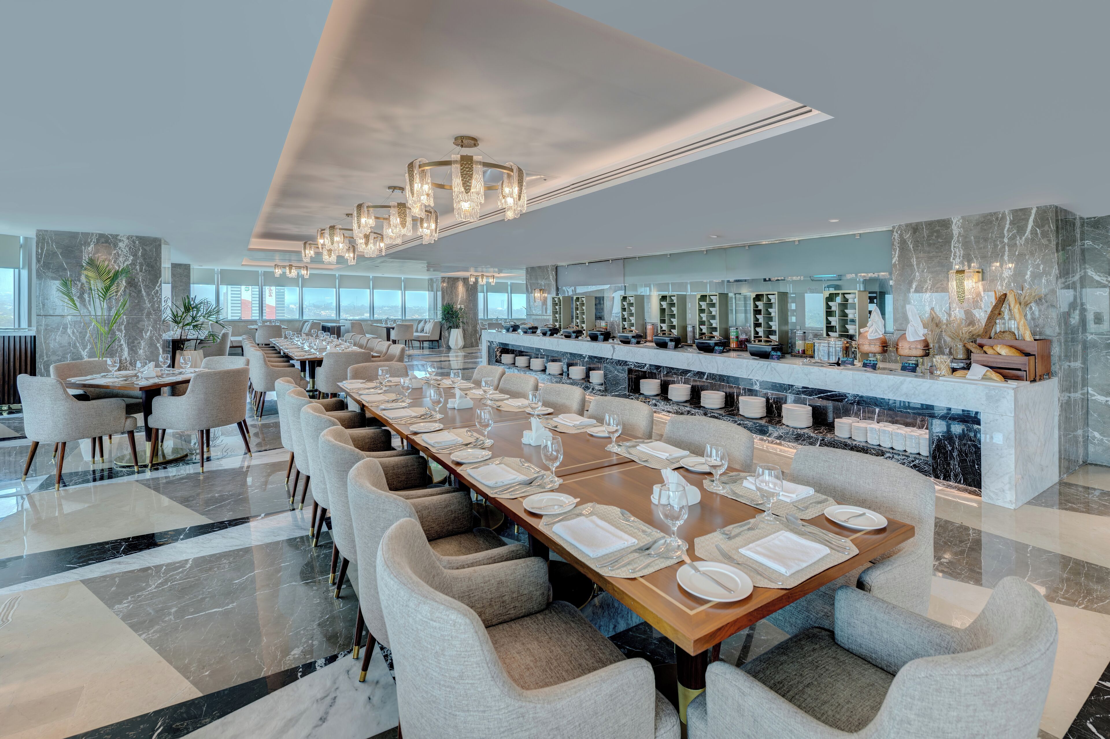 Un comedor moderno con mesas largas preparadas para varios comensales, una encimera de mármol con utensilios de cocina y un amplio ventanal con vistas a la ciudad.