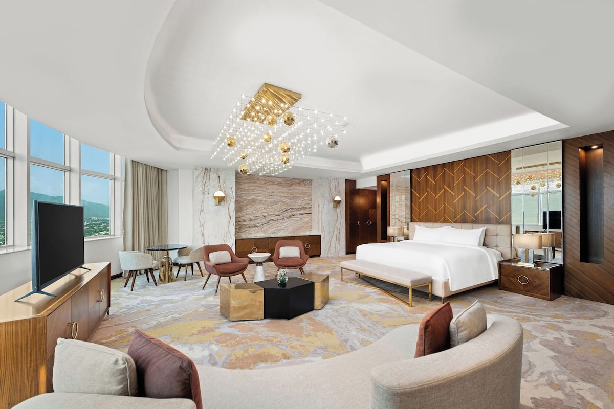 A modern hotel room with a large bed, a seating area with chairs, a TV, and a dining table.