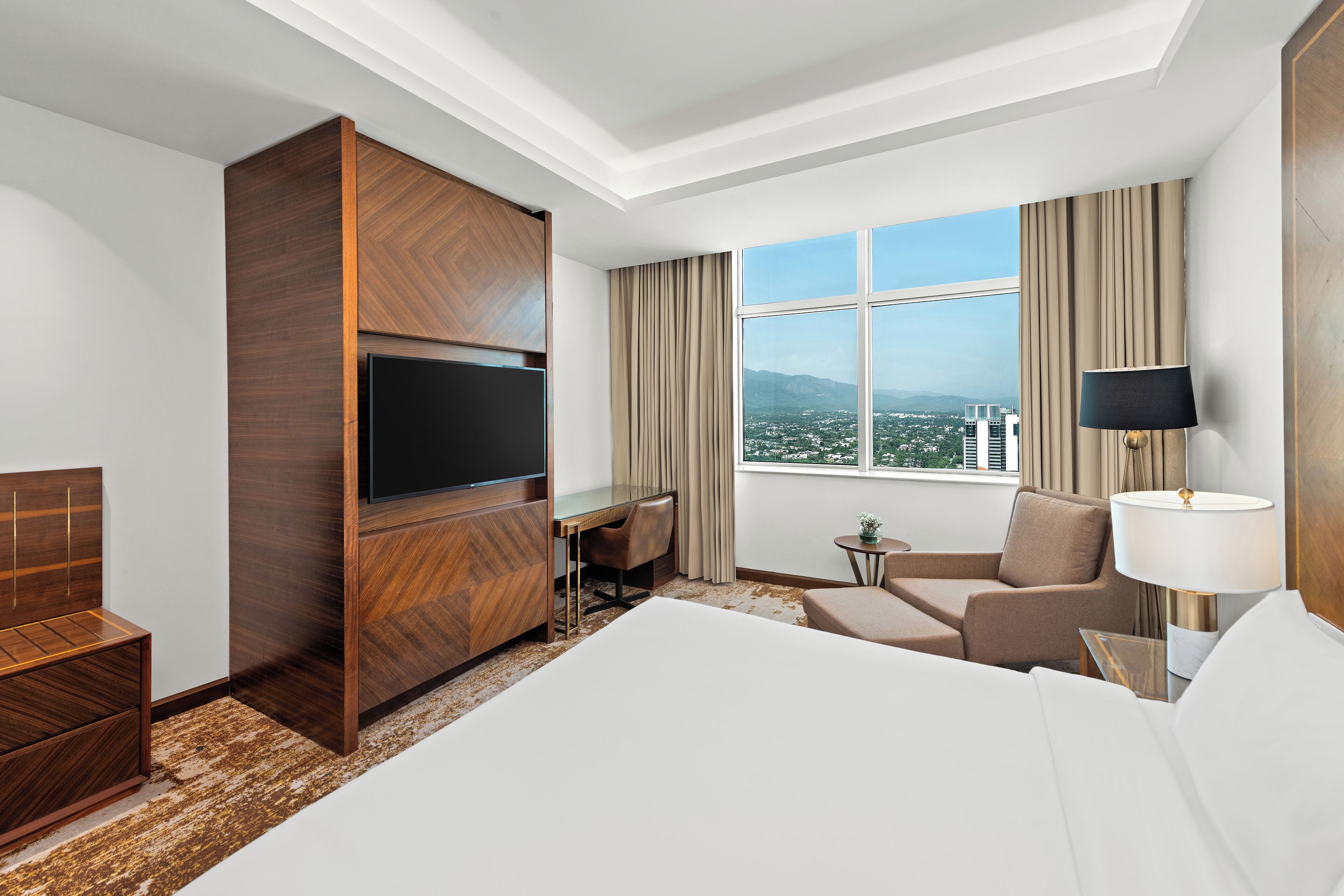 A hotel room with a large bed, a desk, a TV, a chair, and a view of the city.