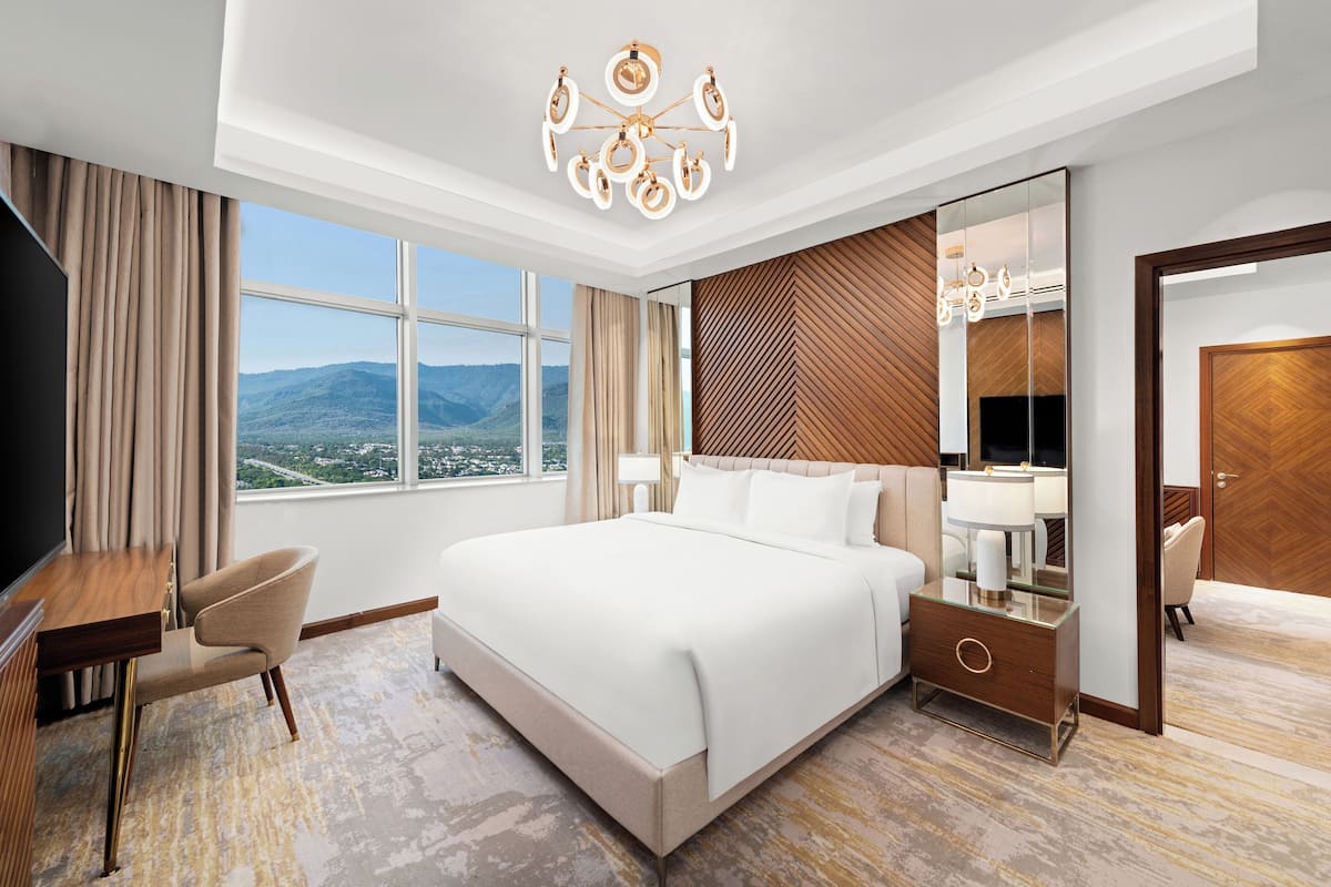 A modern hotel room with a large bed, a desk, and a chair. There is a large window with a mountain view.