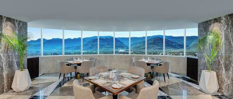 A modern dining area with a large table set for six, surrounded by chairs, with a panoramic mountain view through large windows.