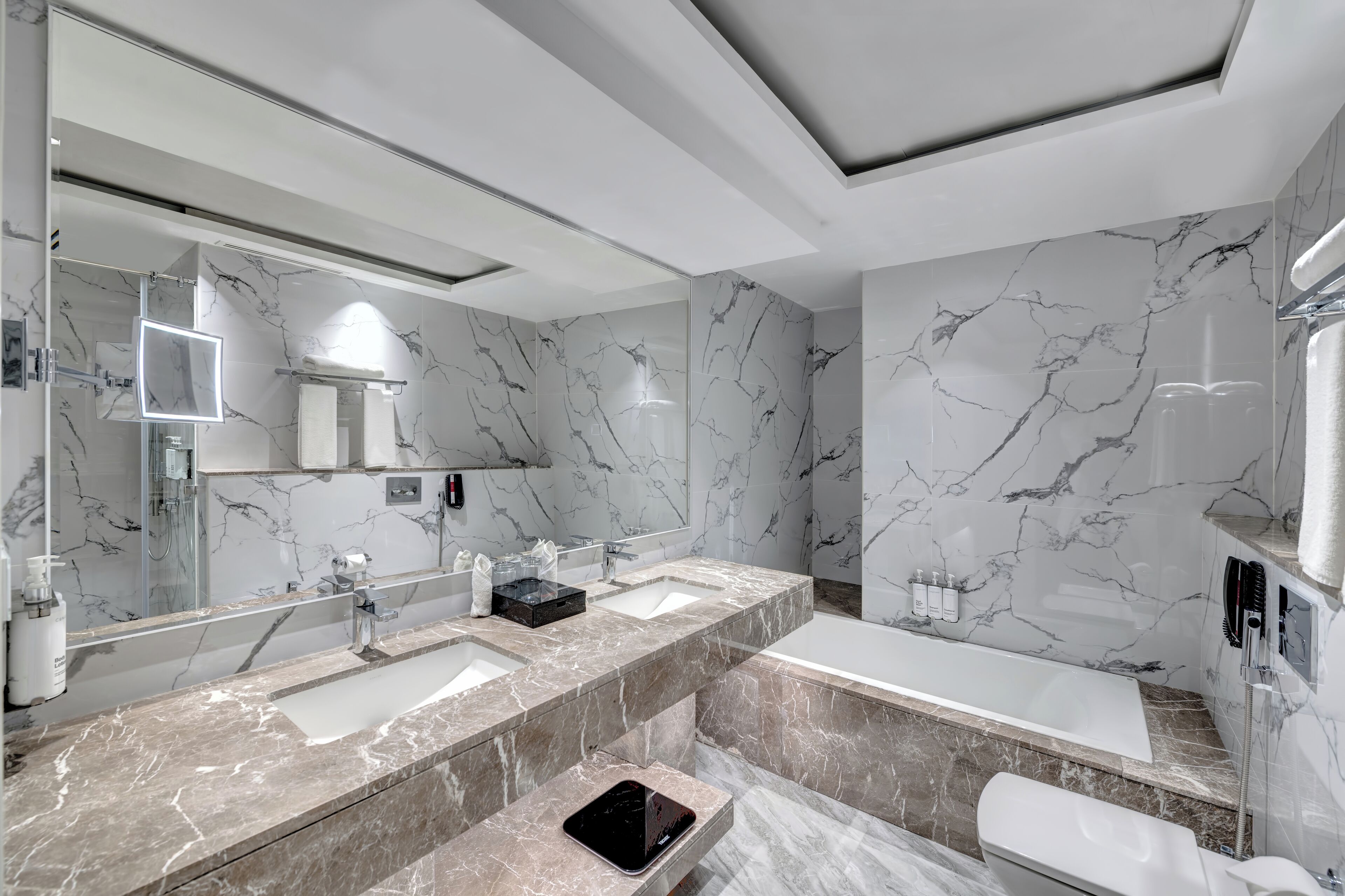 A modern bathroom with marble walls, a large mirror, two sinks, a bathtub, and a handheld shower.