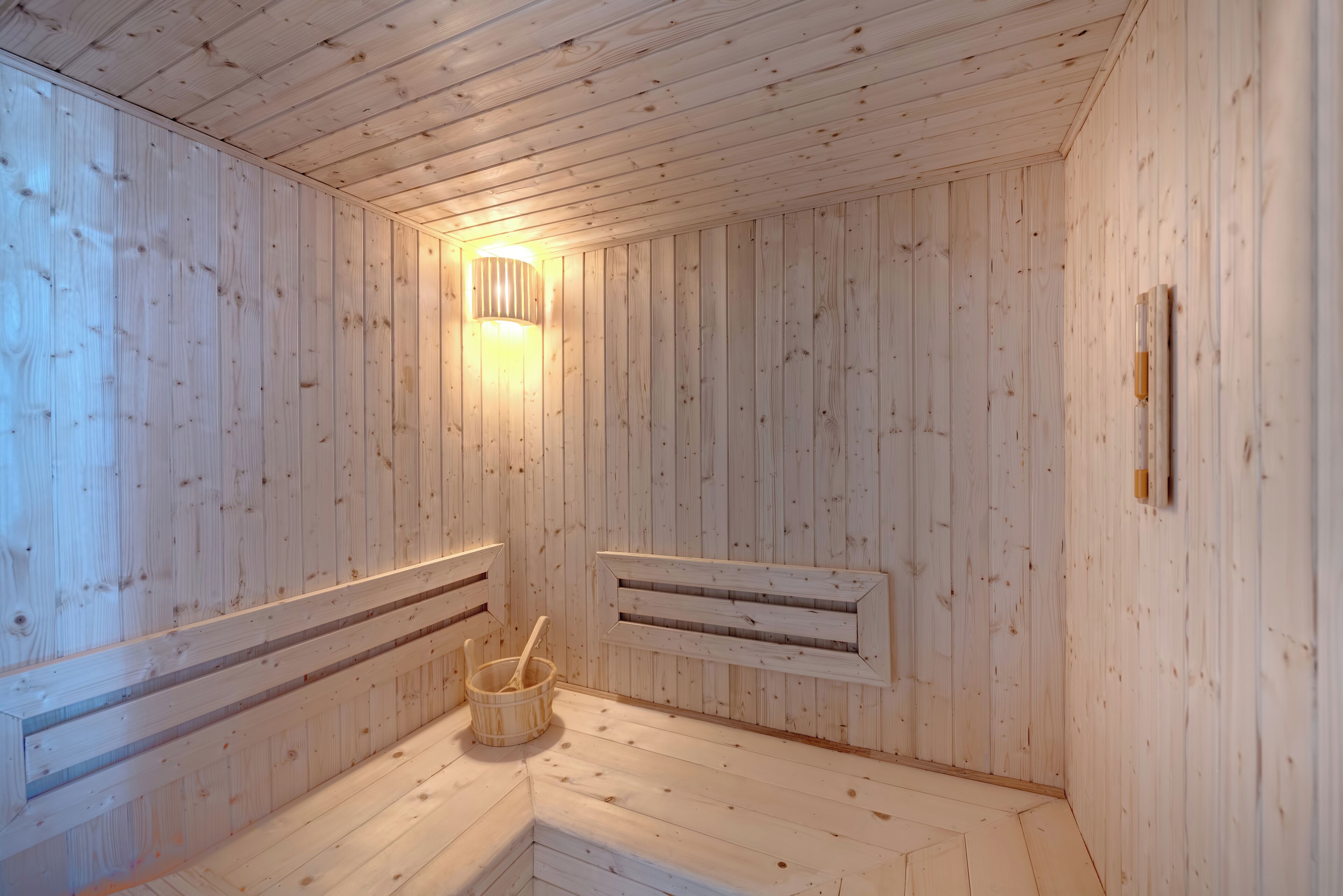 A sauna room with wooden walls, a slanted ceiling, a wall-mounted light fixture, and a wooden bucket with a handle.