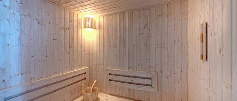 A sauna room with wooden walls, a slanted ceiling, a wall-mounted light fixture, and a wooden bucket with a handle.
