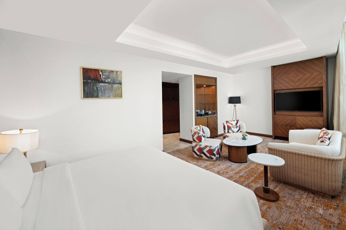 A modern hotel room with a large bed, a sofa, a coffee table, and a TV. There is a painting on the wall and a lamp on the side table.