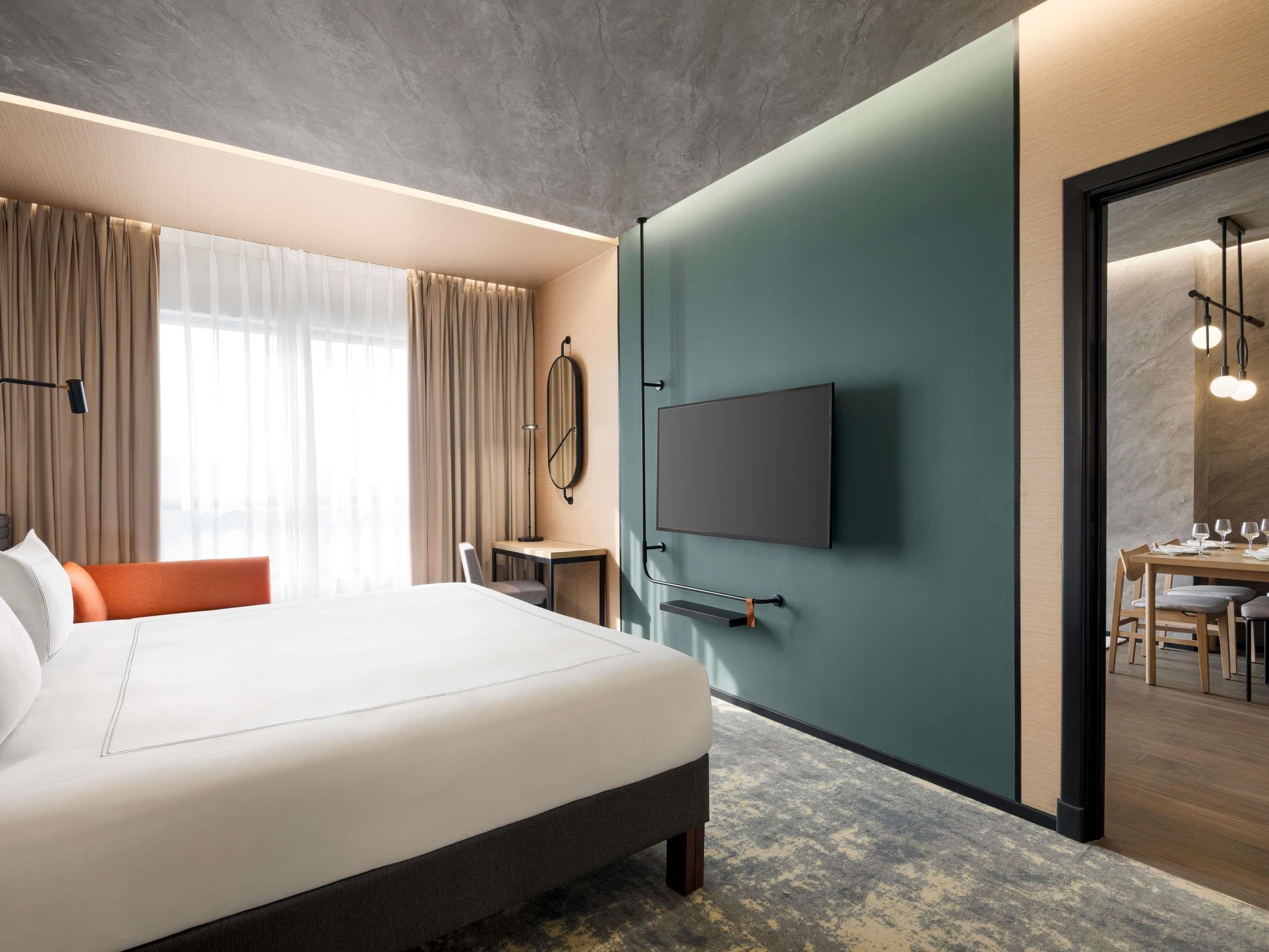 A modern hotel room with a large bed, a desk with a chair, a flat-screen TV mounted on the wall, and a dining area visible through an open door.