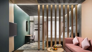 A modern hotel room with a sectional sofa, a small round table with fruit, a desk with a chair, and a TV mounted on the wall. - Novotel Residences Makkah (Makkah)