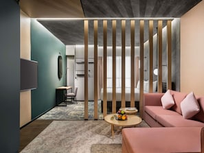 Une chambre d’hôtel moderne avec un canapé d’angle, une petite table ronde sur laquelle sont disposés des fruits, un bureau avec une chaise et un téléviseur fixé au mur.