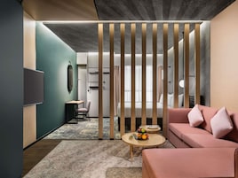 Una habitación de hotel moderna con sofá modular, una mesita redonda con fruta, un escritorio con una silla y un televisor montado en la pared.