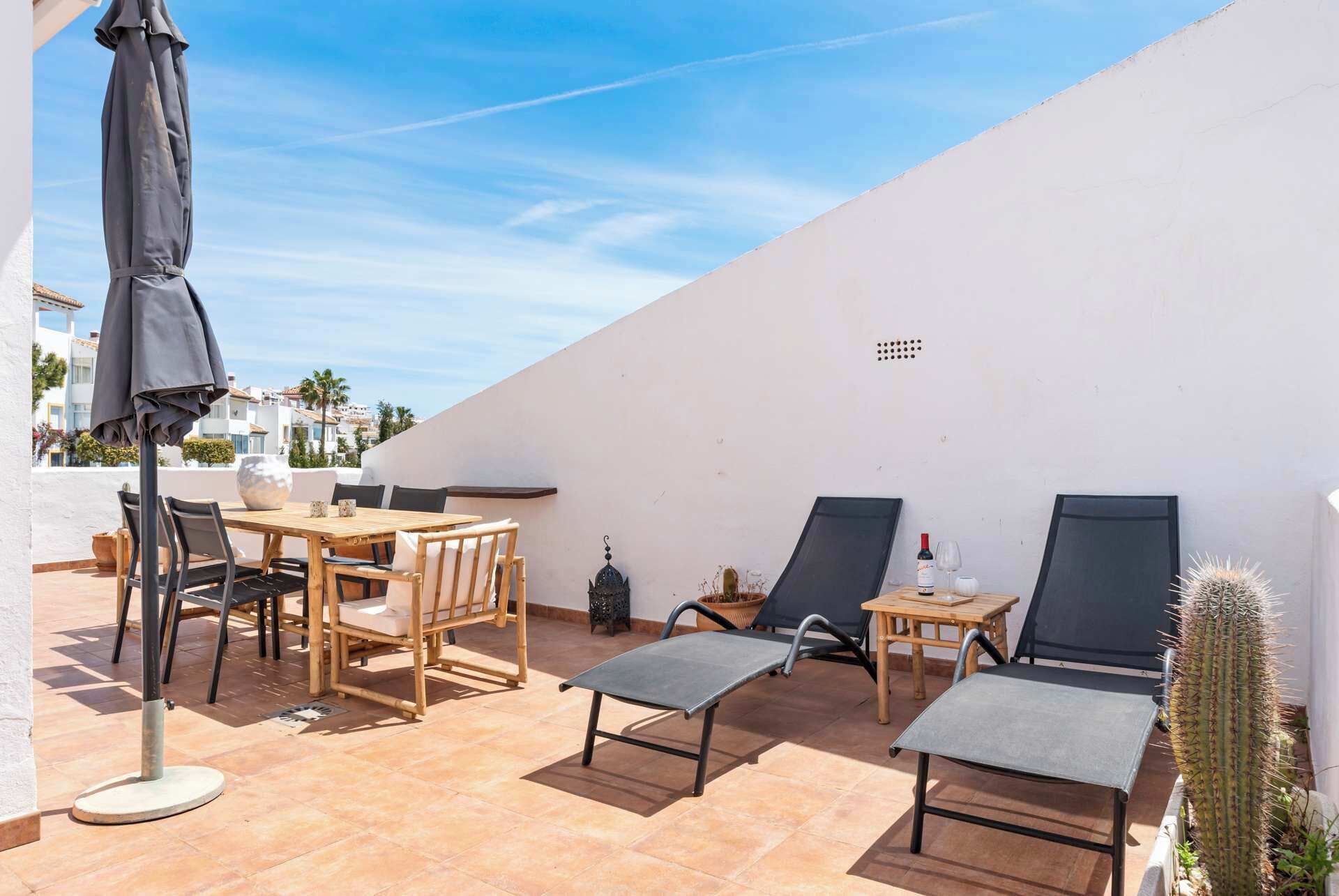 A patio with a table and chairs, a lounge chair, a small table with a bottle and glass, and a cactus plant.