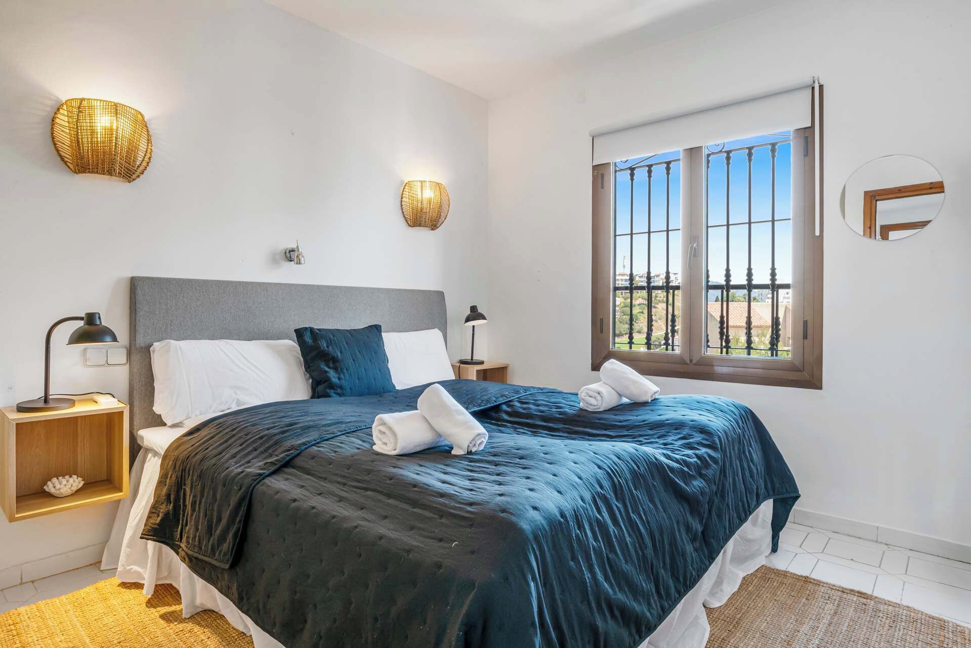 A bedroom with a bed, two bedside lamps, a window with a view, and a wall-mounted light fixture.