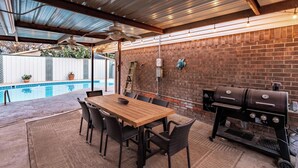 An outdoor patio with a wooden table and chairs, a brick wall, a grill, and a pool in the background.
