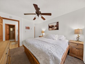 Ein Schlafzimmer mit einem Bett, einem Deckenventilator, einem Nachttisch mit Lampe, einem Holzschrank und einer Tür zu einem anderen Raum.