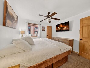 A bedroom with a bed, a ceiling fan, a TV mounted on the wall, and a wooden dresser.