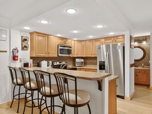 Una cocina moderna con armarios de madera, electrodomésticos de acero inoxidable, una isla central con taburetes de bar e iluminación empotrada en el techo.