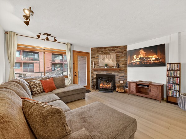 Una sala de estar con chimenea, un conjunto de sofás, una estantería y vista a un edificio.