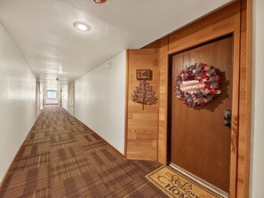 Un pasillo de hotel con una puerta de madera, un felpudo, una corona navideña y un cartel con un número.