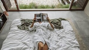 Une personne allongée sur un lit, un livre à la main, dans une pièce dotée d’une grande fenêtre donnant sur un jardin.