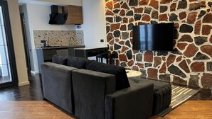 A modern living room with a sectional sofa, a flat-screen TV mounted on a stone wall, and a kitchen in the background. - Garden Lounge Hotel (Yerevan)