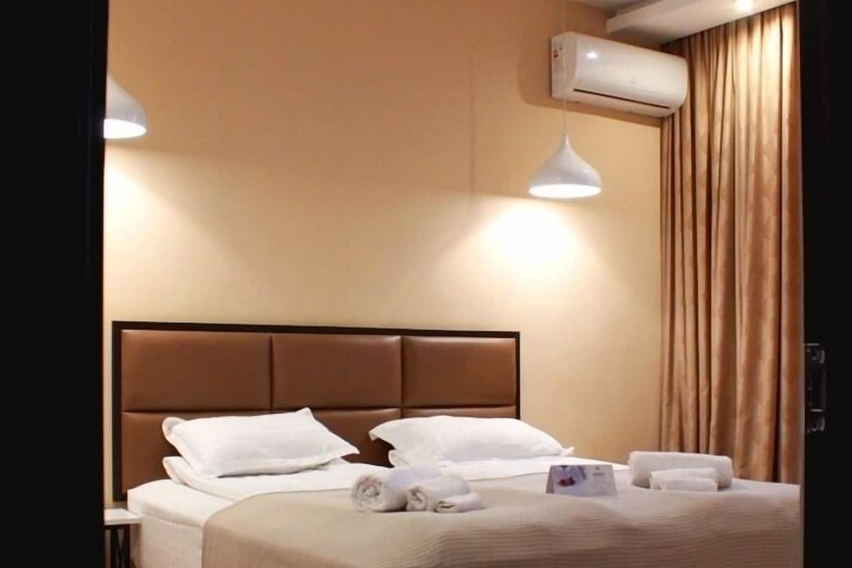 Habitación de hotel con una cama grande, una mesita de noche, aire acondicionado instalado en la pared y una ventana con cortinas.